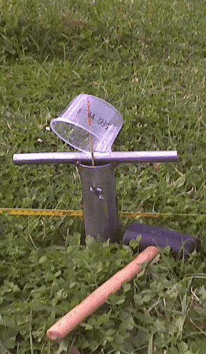 Picture: The photo shows the upper part of a core drill stick for taking soil samples, which is stuck deep in the soil of a meadow. A thin rod protrudes upwards from the device past the cross-handle. A transparent plastic cup hangs above the rod. A yellow folding ruler and a rubber hammer lie on the meadow.