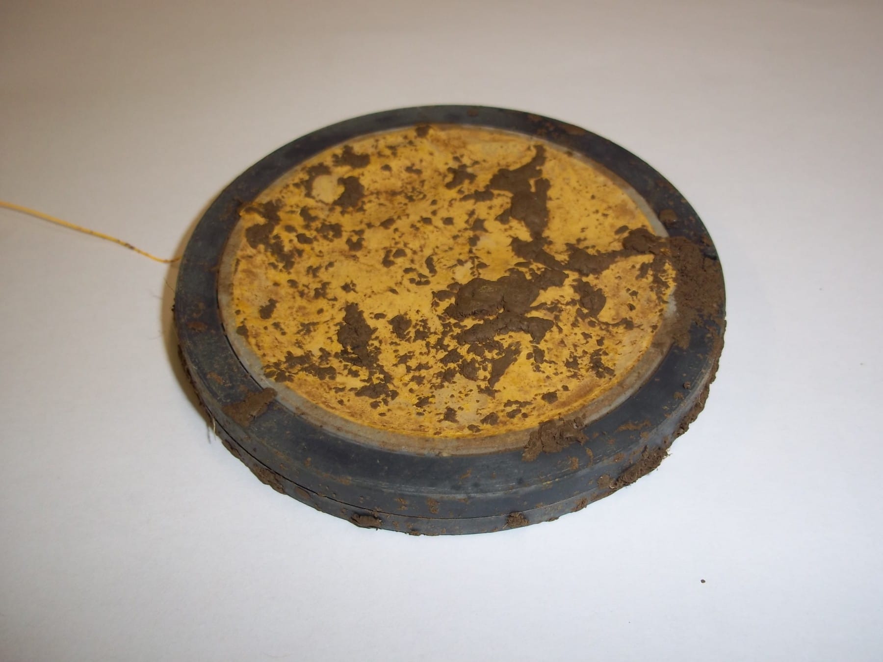 Abbildung: Das Foto zeigt einen auf einem Tisch liegenden flachen runden Mineral-Container nach der Entnahme aus dem Boden. Auf dem Gehäuse und dem Netzbeutel mit der Bodensubstanz befinden sich Reste von Erde.