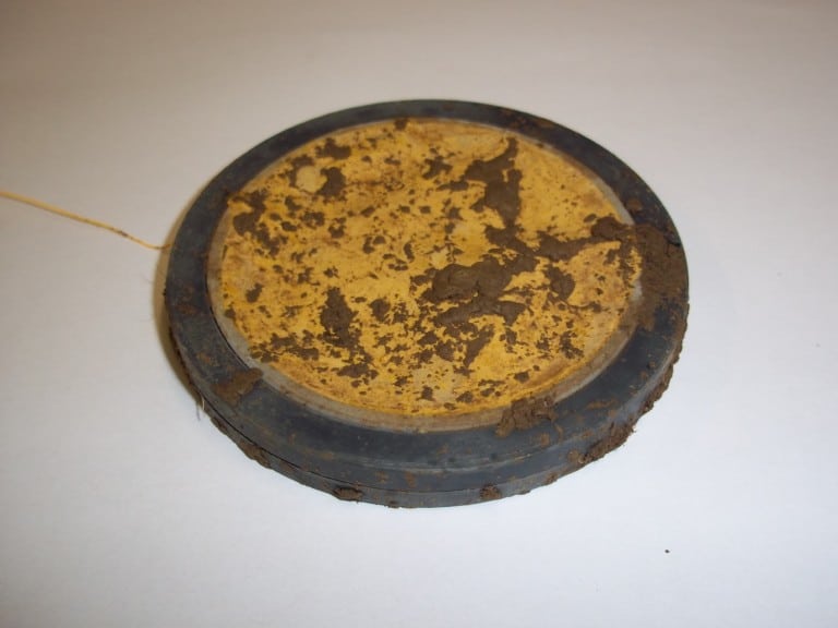 Picture: The photo shows a flat round mineral container lying on a table after removal from the ground. There are remains of soil on the casing and the net bag with the soil substance.