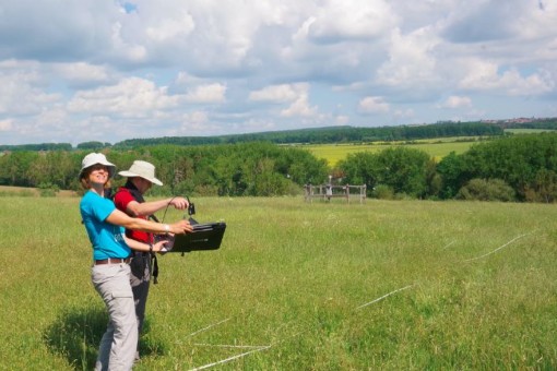 Abbildung: Das Foto zeigt eine ungemähte Wiese, auf der mit weißem Band ein Bereich markiert ist. Im Vordergrund stehen die Projektleiterin Professor Anja Linstädter und der Doktorand Florian Männer, beide mit Hüten zum Schutz vor der Sonne. Frau Linstädter hält auf Höhe der Taille eine schwarze flache Box in ihren Händen. In der Box befindet sich eine Weißreferenz-Platte, über die Herr Männer ein Feld-Spektrometer hält, um es zu kalibrieren.Im Hintergrund des Bilds befinden sich eine umzäunte Klimamess-Station und eine Reihe von Büschen und Bäumen. Am Horizont sind Wiesen und Wälder zu sehen.