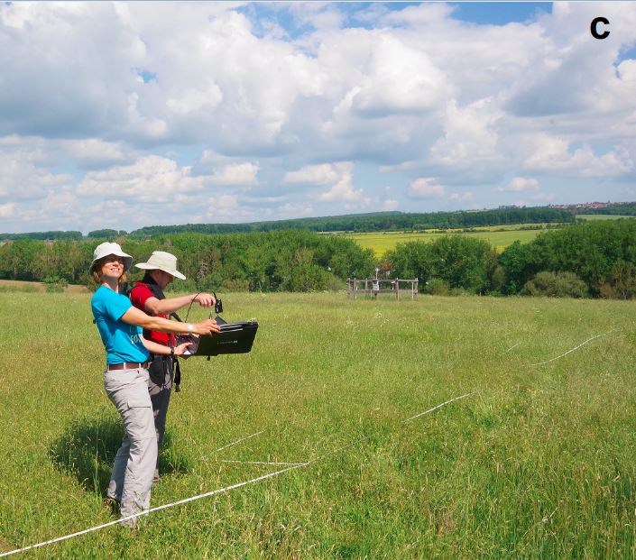 Picture: The photo shows an unmown meadow with an area marked with white tape. In the foreground are project leader Professor Anja Linstädter and PhD student Florian Männer, both wearing hats to protect them from the sun. Ms. Linstädter is holding a black flat box in her hands at waist level. Inside the box is a white reference plate over which Mr. Männer is holding a field spectrometer to calibrate it. In the background of the image is a fenced climate measurement station and a row of bushes and trees. Meadows and forests can be seen on the horizon