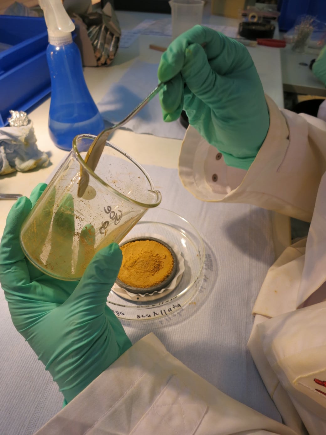 Picture: The photo shows the situation of filling a mineral container. On a work table there is a flat glass bowl containing the mineral container. The round flat container is filled with an orange-brown soil substance. Above the container you can see the arms and hands of a person wearing a lab coat and green latex gloves. The left hand holds a glass beaker while the right hand holds a spoon in the beaker to pick up the remains of the soil substance still in it