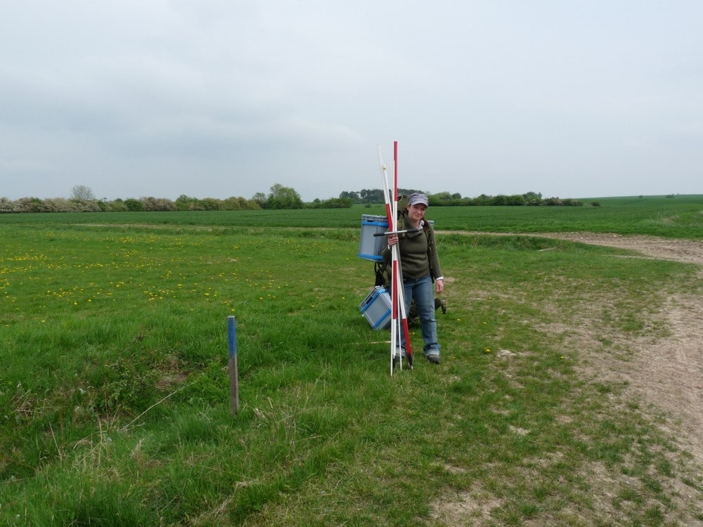 Picture: The photo shows a young female scientist looking into the camera on a meadow in the Hainich-Dün-Exploratory. In her left hand the woman is holding an earth drill stick for taking soil samples as well as two thin white sticks and two red and white striped escape sticks. On her back the woman is carrying two grey plastic boxes containing soil samples.