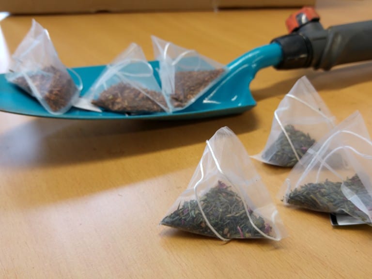 Picture: The photo shows a table in a room with an orange-brown top with a wood grain. A turquoise hand shovel lies on the table. Three tea bags filled with tea lie on the shovel and on the table top in front of it.